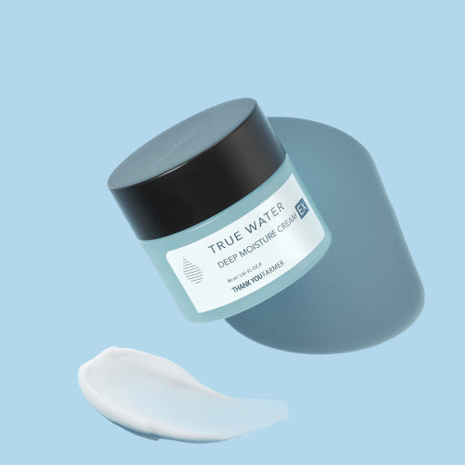 A jar of THANK YOU FARMER True Water Deep Moisture Cream EX with a small amount of cream placed beside it, on a light blue background.