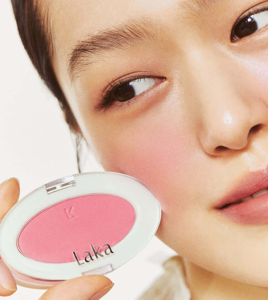 Model applying Laka Love Silk Blush in warm pink shade, showcasing luminous pearls for a transparent complexion.